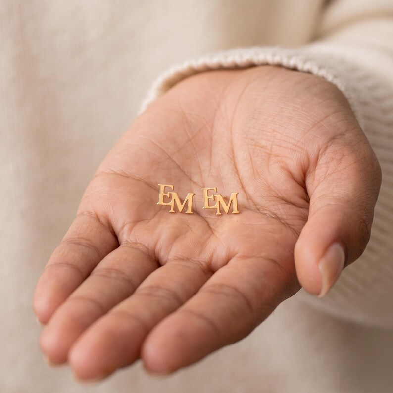 Hand holding a pair of two letter monogram studs in an open palm, showing both gold EM earrings side by side to give a sense of their petite everyday size.