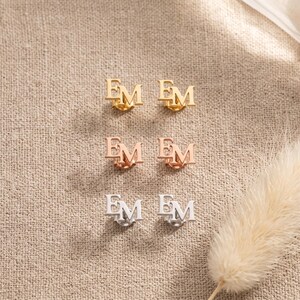 Interlocking initial stud earrings in gold, rose gold and silver plating laid in rows on linen with bunny tail grass, showing the three plating color choices.
