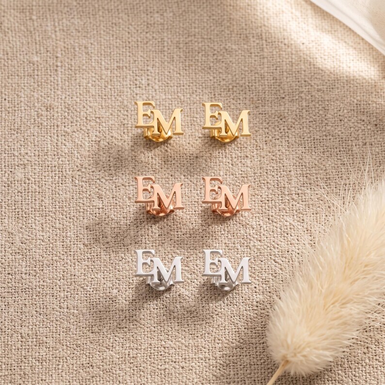 Interlocking initial stud earrings in gold, rose gold and silver plating laid in rows on linen with bunny tail grass, showing the three plating color choices.