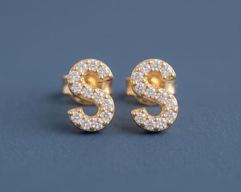 May include: A pair of gold stud earrings shaped like the letter S, each featuring a series of small, clear, round-cut stones set along the surface of the letter. The earrings have a polished gold finish and include standard post-style backings.