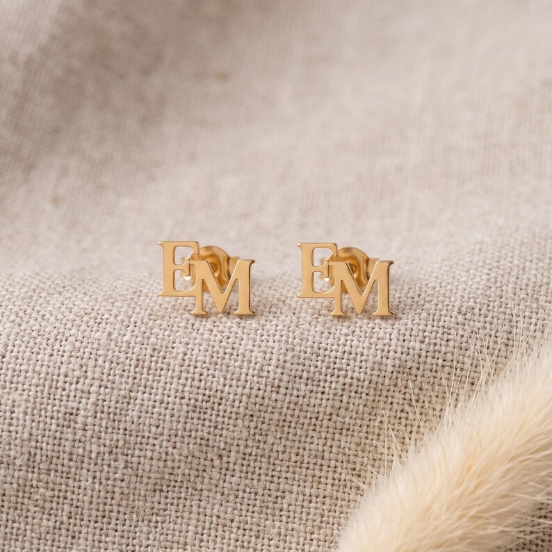 Pair of gold monogram earrings resting on beige linen with a bunny tail grass stem, showing the interlocking E and M letters cut together as one tiny stud each.