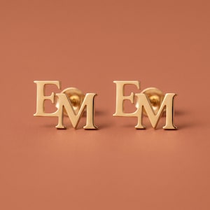 May include: A pair of gold-toned stud earrings, each featuring the capital letters EM in a serif font. The earrings have a polished, smooth finish and include standard post-style backings for secure attachment to the earlobe.