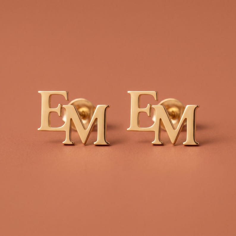 Close-up of interlocking initial stud earrings on a terracotta background, showing a pair of gold monogram studs with serif letters E and M woven together as one piece.