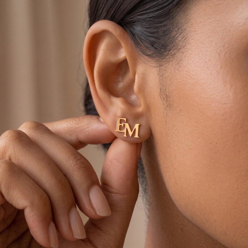 Fingers touching a gold initial stud earring on the earlobe, showing the dainty EM monogram up close and how neatly the double initial stud sits when worn.