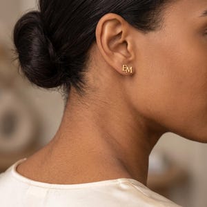 Side view of a woman wearing a double initial stud earring with her hair in a low bun, showing the small gold EM monogram sitting neatly on the earlobe.