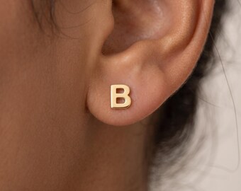 May include: A small, gold-coloured stud earring shaped like the capital letter B. The earring features a smooth, polished surface and a simple, block-style font design. It is positioned directly on the earlobe, showcasing a clean and metallic finish.