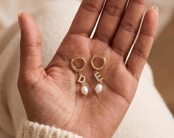 May include: A pair of gold hoop earrings featuring dangling letter charms D and E, each encrusted with small clear stones. Below each letter charm hangs a single white oval-shaped pearl. The earrings are crafted from a polished gold-tone metal.