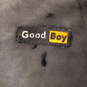 May include: A dark gray, soft-textured fabric featuring a rectangular black label stitched onto the surface. The label displays the white text 'Good' followed by the word 'Boy' in black lettering inside a bright yellow rectangular box.