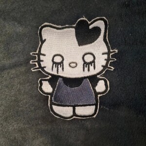 May include: An embroidered patch featuring a stylized white cat character with a black heart on its head and black dripping eyes. The character wears a dark gray dress and has black whiskers on both sides of its face, outlined in black thread.