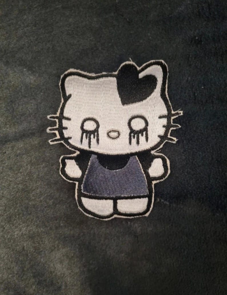 May include: An embroidered patch featuring a stylized white cat character with a black heart on its head and black dripping eyes. The character wears a dark gray dress and has black whiskers on both sides of its face, outlined in black thread.