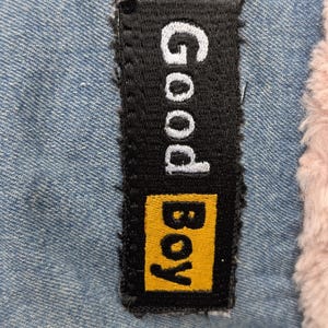 May include: A rectangular black embroidered patch featuring the white text Good and the yellow text Boy, stitched onto a light blue denim fabric next to a section of soft, light pink faux fur material with a textured, fuzzy appearance.