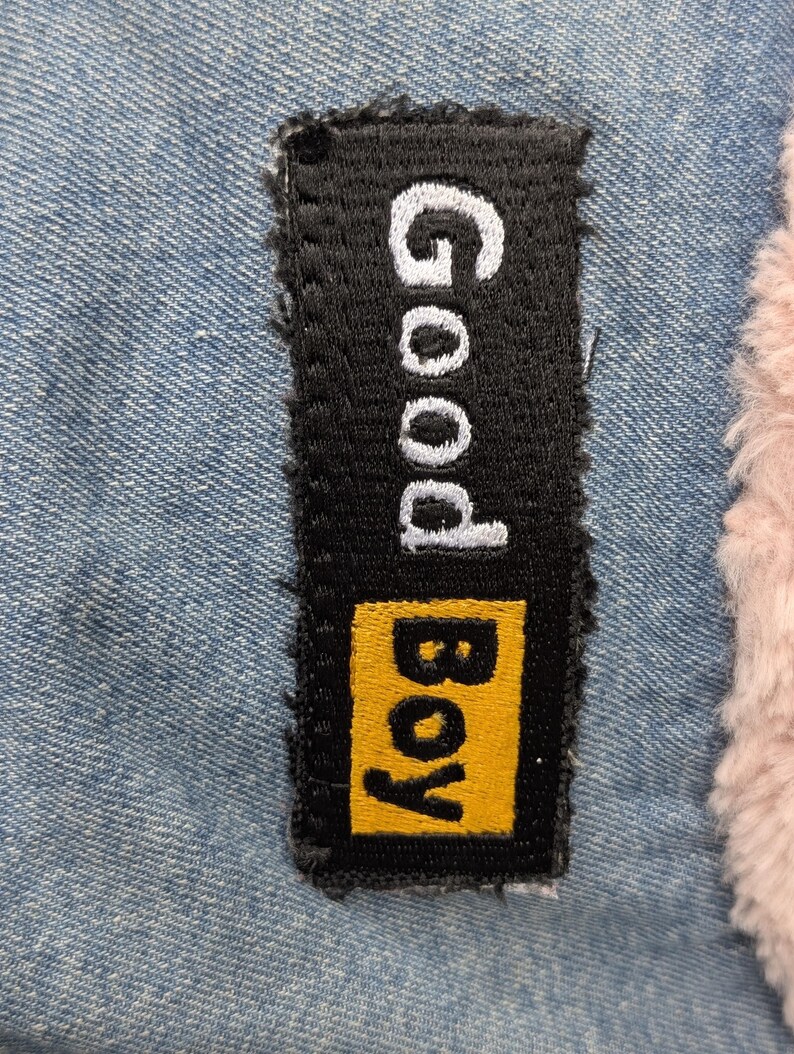 May include: A rectangular black embroidered patch featuring the white text Good and the yellow text Boy, stitched onto a light blue denim fabric next to a section of soft, light pink faux fur material with a textured, fuzzy appearance.