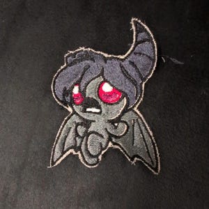 Puede incluir: Un parche bordado que muestra una criatura gris oscuro estilizada con un cuerno curvado, grandes ojos rojos brillantes y pequeñas alas de murciélago. El personaje tiene una cabeza redondeada, una boca pequeña y un estilo oscuro.
