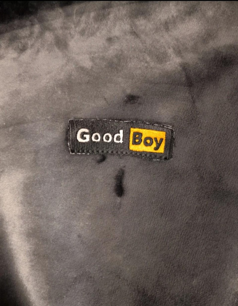 May include: A dark gray, soft-textured fabric featuring a rectangular black label stitched onto the surface. The label displays the white text 'Good' followed by the word 'Boy' in black lettering inside a bright yellow rectangular box.