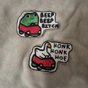 Könnte beinhalten: Zwei bestickte Aufnäher mit weißem Rand. Der obere Aufnäher zeigt einen grünen Frosch in einem roten Auto mit dem Text BEEP BEEP BITCH. Der untere Aufnäher zeigt eine weiße Gans in einem roten Auto mit dem schwarzen Text HONK HONK HOE.