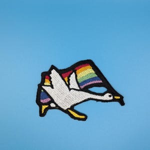 Könnte beinhalten: Ein gestickter Aufnäher mit einer weißen Gans im Flug, die eine regenbogenfarbene Flagge trägt. Das Motiv ist mit dickem schwarzem Garn umrandet, die Gans hat einen gelben Schnabel und gelbe Füße, vor einem hellblauen Hintergrund.
