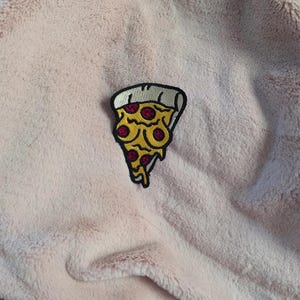 May include: An embroidered patch shaped like a slice of pizza with yellow cheese, red pepperoni, and a white crust, featuring a black outline. The patch is attached to a soft, light pink, fuzzy fabric with a plush, textured surface.