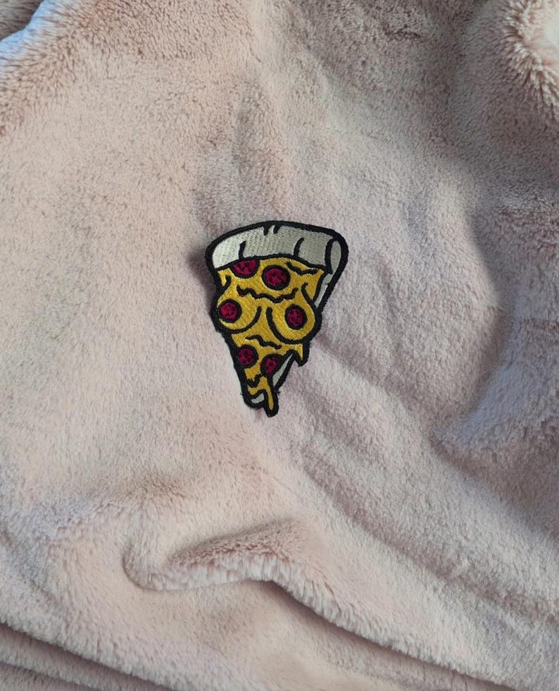May include: An embroidered patch shaped like a slice of pizza with yellow cheese, red pepperoni, and a white crust, featuring a black outline. The patch is attached to a soft, light pink, fuzzy fabric with a plush, textured surface.
