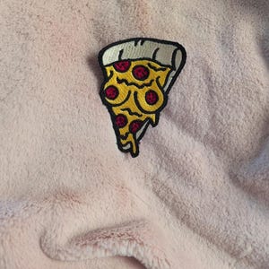 May include: An embroidered patch shaped like a slice of pizza with a thick white crust, yellow melted cheese, and red pepperoni circles. The patch features a bold black outline and is positioned on a soft, light pink, textured fabric surface.