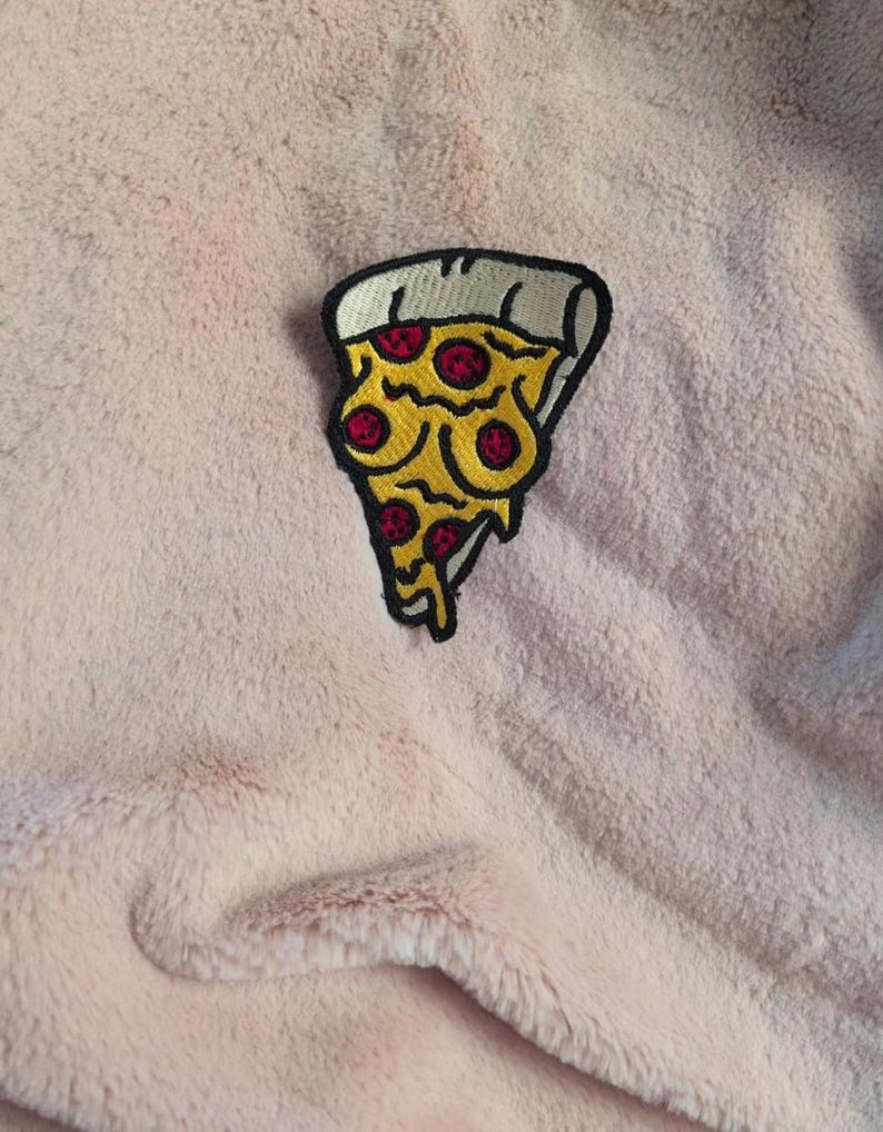 May include: An embroidered patch shaped like a slice of pizza with a thick white crust, yellow melted cheese, and red pepperoni circles. The patch features a bold black outline and is positioned on a soft, light pink, textured fabric surface.