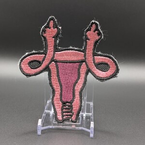 May include: An embroidered patch featuring a stylized anatomical uterus in shades of pink and dark maroon with black outlines. The fallopian tubes extend upward and terminate in shapes resembling hands giving the middle finger gesture.