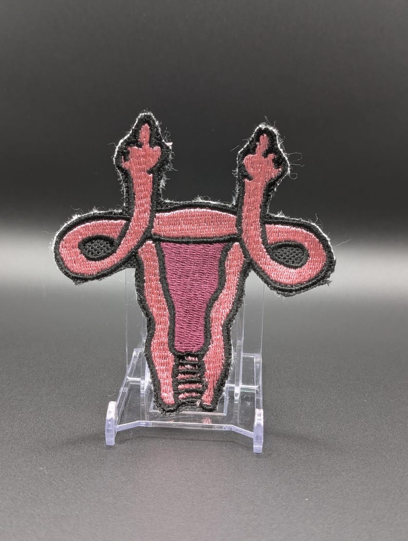 May include: An embroidered patch featuring a stylized anatomical uterus in shades of pink and dark maroon with black outlines. The fallopian tubes extend upward and terminate in shapes resembling hands giving the middle finger gesture.