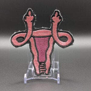 May include: An embroidered patch shaped like a uterus in shades of pink and dark purple with a black border. The fallopian tubes extend upward and outward, each ending in a hand gesture with the middle finger raised in a defiant manner.
