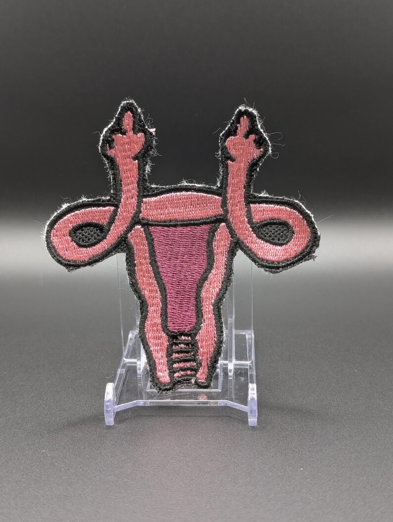 May include: An embroidered patch shaped like a uterus in shades of pink and dark purple with a black border. The fallopian tubes extend upward and outward, each ending in a hand gesture with the middle finger raised in a defiant manner.