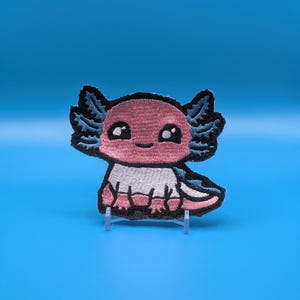 May include: An embroidered patch featuring a cartoon axolotl with a pink head, white body, and dark blue gills. The design has a thick black outline, large black eyes with white highlights, and a small smile, all rendered in detailed thread work.