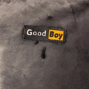 May include: A dark gray, soft-textured fabric surface featuring a rectangular black fabric label stitched onto it. The label displays the white text "Good" followed by the word "Boy" in black lettering inside a bright yellow rectangular box.