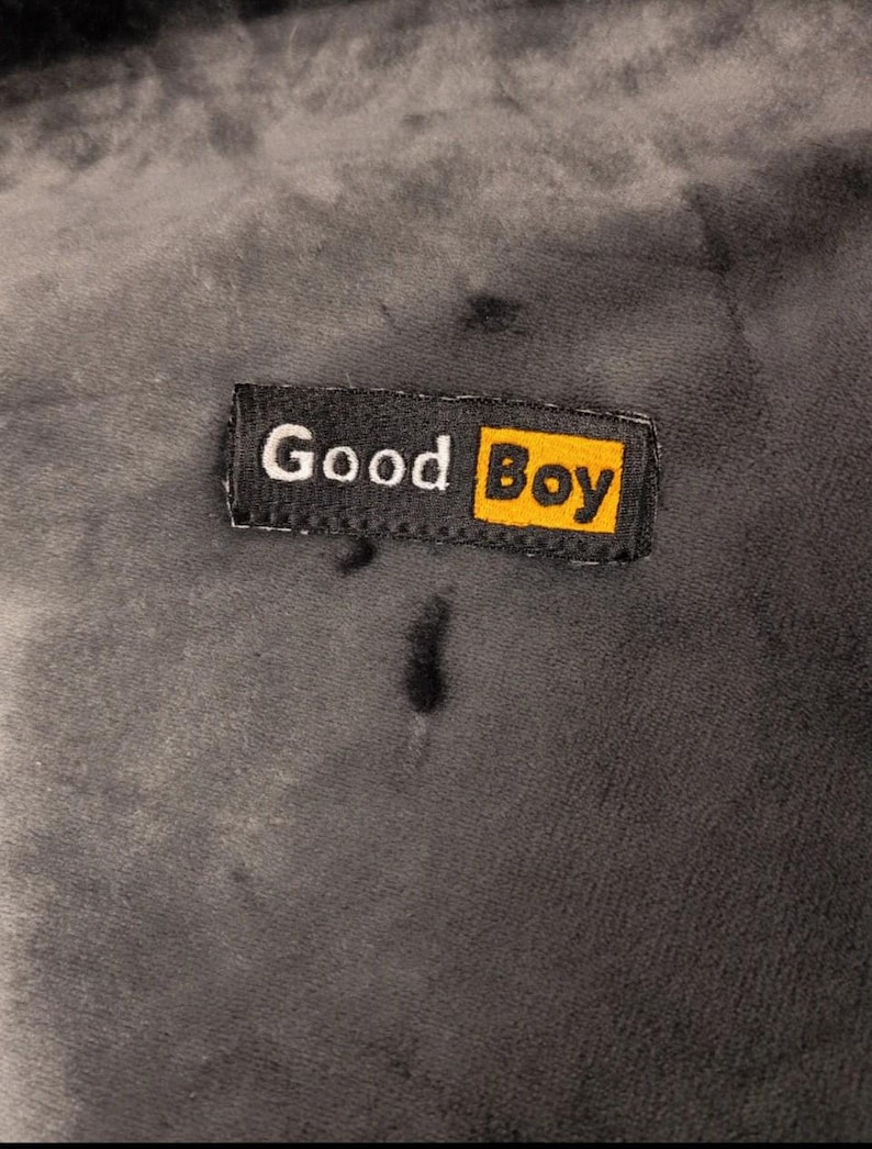 May include: A dark gray, soft-textured fabric surface featuring a rectangular black fabric label stitched onto it. The label displays the white text "Good" followed by the word "Boy" in black lettering inside a bright yellow rectangular box.