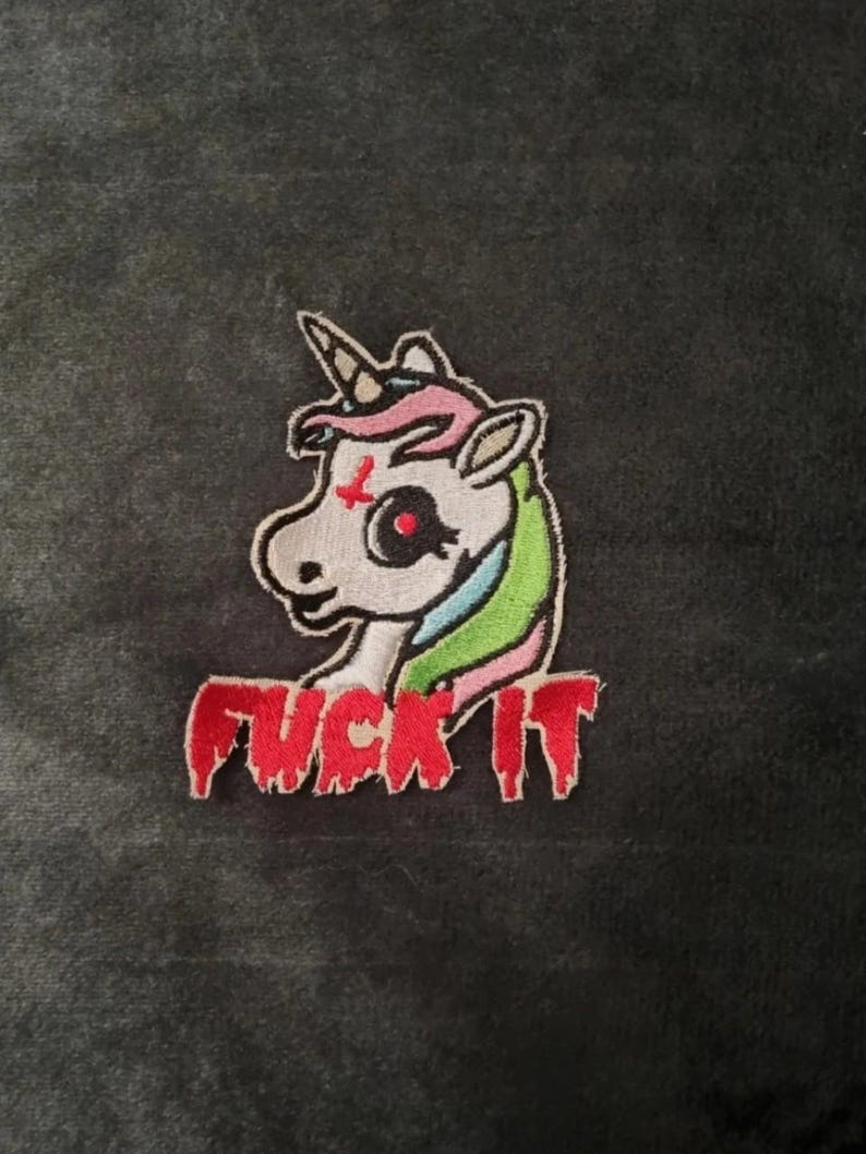 May include: An embroidered patch featuring a white unicorn head with a red inverted cross on its forehead, a black eye, and a multicolored mane in pink, blue, and green. Below the unicorn, the text FUCK IT is stitched in a dripping red font.