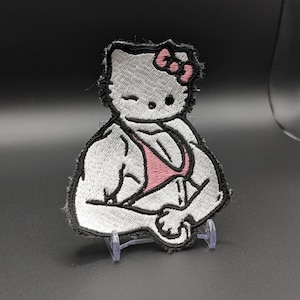 Könnte beinhalten: Ein gestickter Aufnäher, der eine weiße Katzenfigur mit einer rosa Schleife zeigt, die ein rosa Bikini-Oberteil trägt und muskulöse Arme in einer Beugepose präsentiert. Das Design ist mit schwarzem Garn auf weißem Grund umrandet.