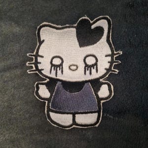 May include: An embroidered patch featuring a white cat character with a black heart on its head, black whiskers, and black tear-like markings under its eyes. The character wears a dark gray dress and has a thick black outline around its entire shape.