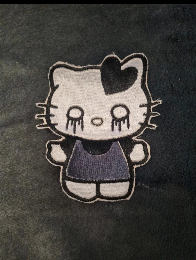 May include: An embroidered patch featuring a white cat character with a black heart on its head, black whiskers, and black tear-like markings under its eyes. The character wears a dark gray dress and has a thick black outline around its entire shape.