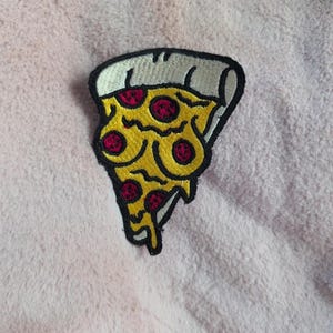 May include: An embroidered patch shaped like a slice of pizza with a thick black border. The pizza features a white crust, yellow melted cheese, and five circular red pepperoni toppings. The design has a stylized, cartoon-like appearance.