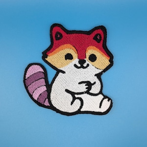 Può includere: Una toppa ricamata raffigurante un procione in stile cartoon con fronte rossa, guance color sabbia e corpo bianco. Il procione ha naso e occhi neri e una coda a strisce con sfumature di viola alternate. Il design è rifinito con un bordo nero.