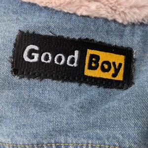 May include: A light blue denim jacket featuring a rectangular black patch with the white and yellow embroidered text Good Boy. The jacket is lined with a soft, light pink faux fur material visible along the top edge of the garment.