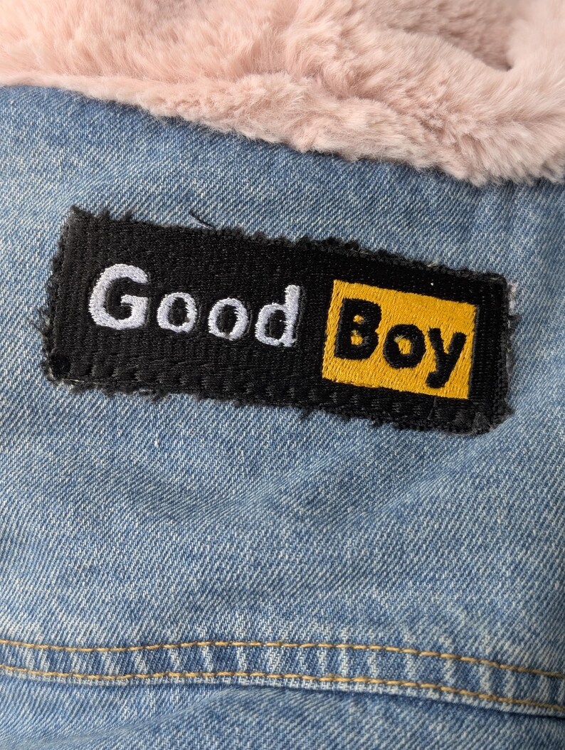 May include: A light blue denim jacket featuring a rectangular black patch with the white and yellow embroidered text Good Boy. The jacket is lined with a soft, light pink faux fur material visible along the top edge of the garment.