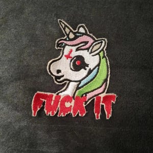May include: An embroidered patch featuring a white unicorn with a red inverted cross on its forehead, a single horn, and a multicolored mane. Below the unicorn, the text FUCK IT is stitched in red with a dripping, liquid-like effect.