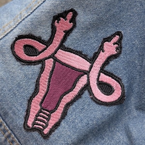 May include: An embroidered patch featuring a stylized pink and dark purple uterus shape with fallopian tubes that extend upward and end in hands making a middle finger gesture, all outlined in black thread on a light blue denim fabric background.
