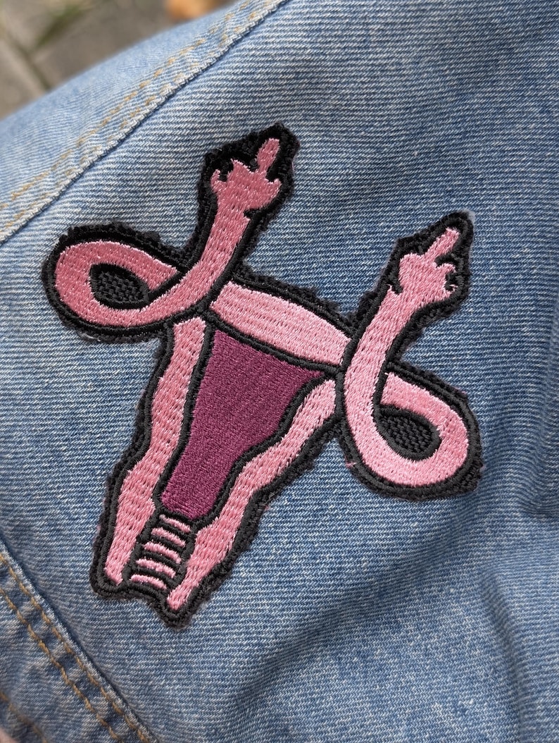 May include: An embroidered patch featuring a stylized pink and dark purple uterus shape with fallopian tubes that extend upward and end in hands making a middle finger gesture, all outlined in black thread on a light blue denim fabric background.