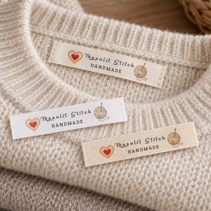 Könnte beinhalten: Drei rechteckige Textiletiketten für Kleidung mit dem Text Moonlit Stitch HANDMADE, einem kleinen roten Herzsymbol und einer braunen Wollknäuelgrafik, platziert auf einem Stapel gefalteter cremefarbener und grauer Strickpullover.
