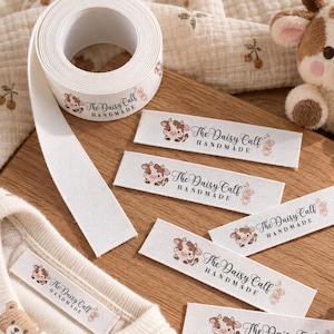 May include: A roll and several individual white fabric labels featuring a brown and white cartoon calf illustration and the text The Daisy Calf HANDMADE. The labels are displayed alongside a plush calf toy and a cream-colored garment.