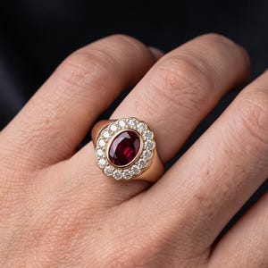 Puede incluir: Un anillo de oro con una piedra preciosa roja oscura de corte ovalado en el centro, rodeada por un halo de pequeñas piedras blancas redondas. El anillo se lleva junto a una delicada pulsera de cadena dorada con una piedra verde y cuentas rojas.