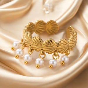 Puede incluir: Un anillo abierto de tono dorado con una banda compuesta por motivos de conchas repetidos. Seis pequeñas perlas blancas redondas cuelgan del borde inferior de la banda de conchas mediante pequeñas anillas metálicas doradas.