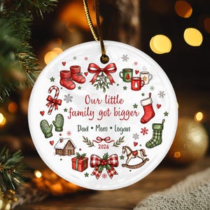 May include: A round white ceramic Christmas ornament featuring a gold hanging cord. The design includes red and green holiday illustrations, such as stockings, mittens, a cabin, and a rocking horse. Text reads: Our little family got bigger, Dad, Mom, Logan, 2026.