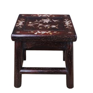 May include: A dark brown wooden stool with a square top featuring inlaid mother-of-pearl floral and bird designs. The stool has four sturdy legs connected by horizontal support bars on all sides, showcasing a visible wood grain texture throughout.