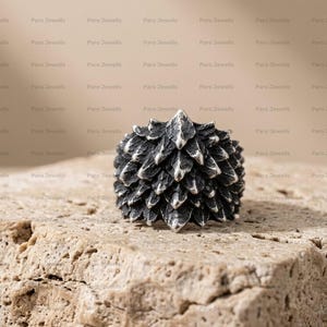 May include: A silver ring featuring a textured, layered scale design with a dark, oxidized finish. The surface of the ring is composed of overlapping, pointed segments that create a dimensional, rugged appearance across the entire band.