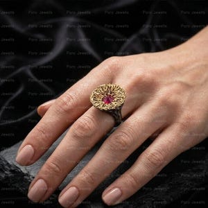 May include: A ring featuring a textured, gold-toned, organic-shaped top with a deep pink, faceted, round gemstone set in the center. The band of the ring is dark, with a rough, uneven surface texture that complements the intricate, coral-like design.